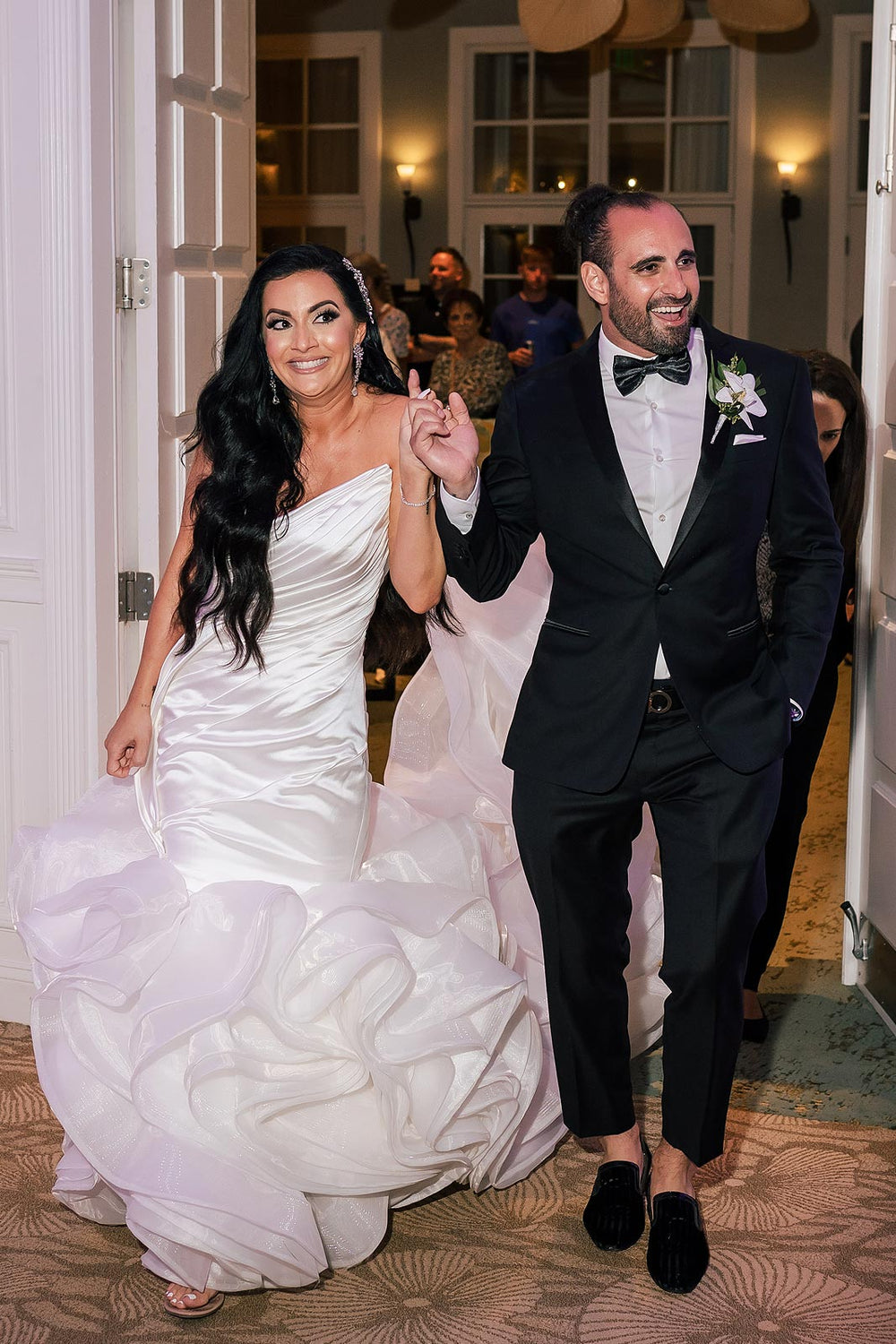 Bride and groom making a joyful grand entrance into the reception hall in the Chelsea mermaid wedding dress by Angela Kim Couture, full gown and voluminous organza ruffle cathedral train visible.
