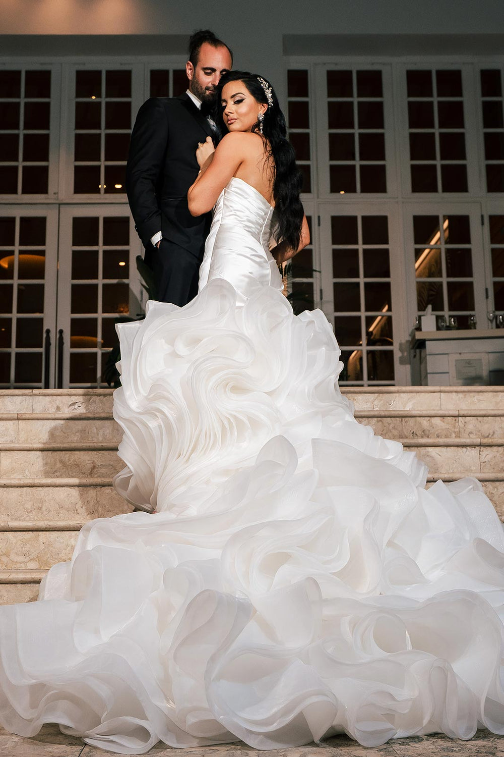 Bride and groom evening portrait at the wedding venue with the Chelsea satin mermaid wedding dress by Angela Kim Couture, groom embracing bride from behind as the layered organza ruffle train pools at the base