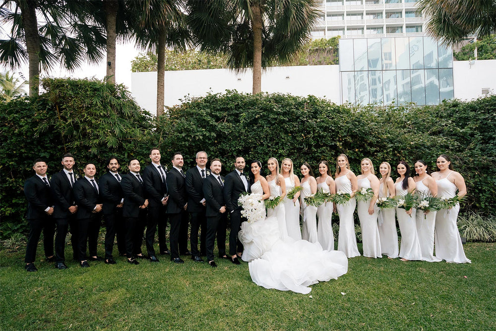 Bride wearing the Chelsea wedding dress by Angela Kim Couture standing with a companion in front of a lush ivy wall, bride holding a white orchid bouquet and wearing large crystal drop earrings with asymmetric satin bodice