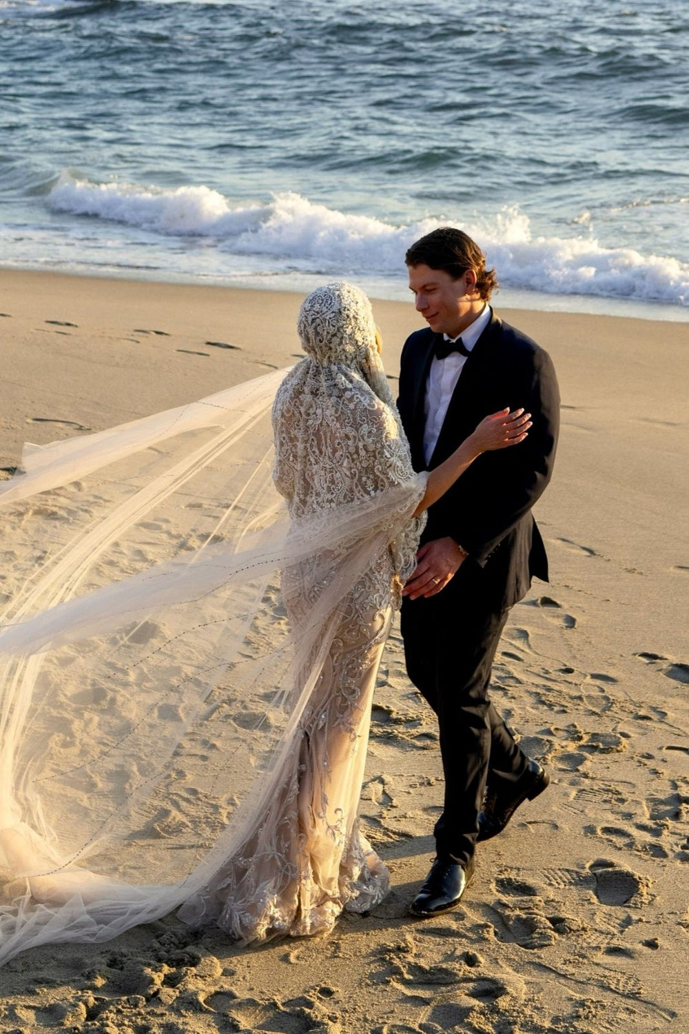 Bride and groom walking on the beach with the Gigi couture lace veil flowing dramatically in the wind