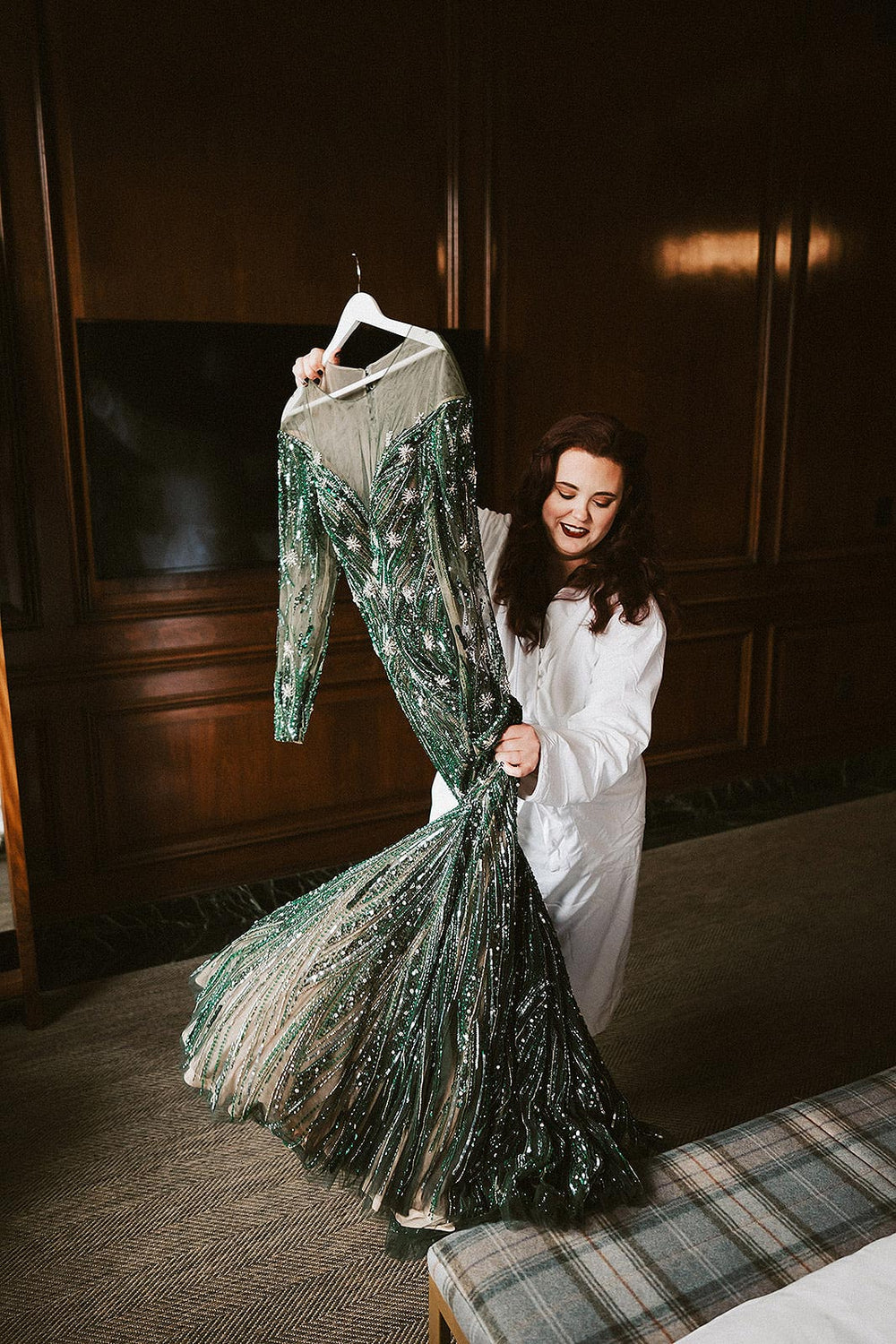 Bride in a white robe holding up her forest green beaded wedding gown on a hanger and gazing at it with excitement — the Jessica custom hand-beaded green wedding gown by Angela Kim Couture displayed in full length showing the trumpet silhouette and crystalline beadwork