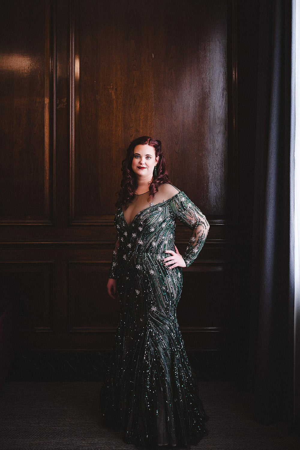Full-length bridal portrait of bride posing with hand on hip in front of dark wood paneling, wearing the Jessica forest green trumpet wedding gown by Angela Kim Couture — the star and stripe crystal-beaded pattern across the fitted silhouette clearly visible