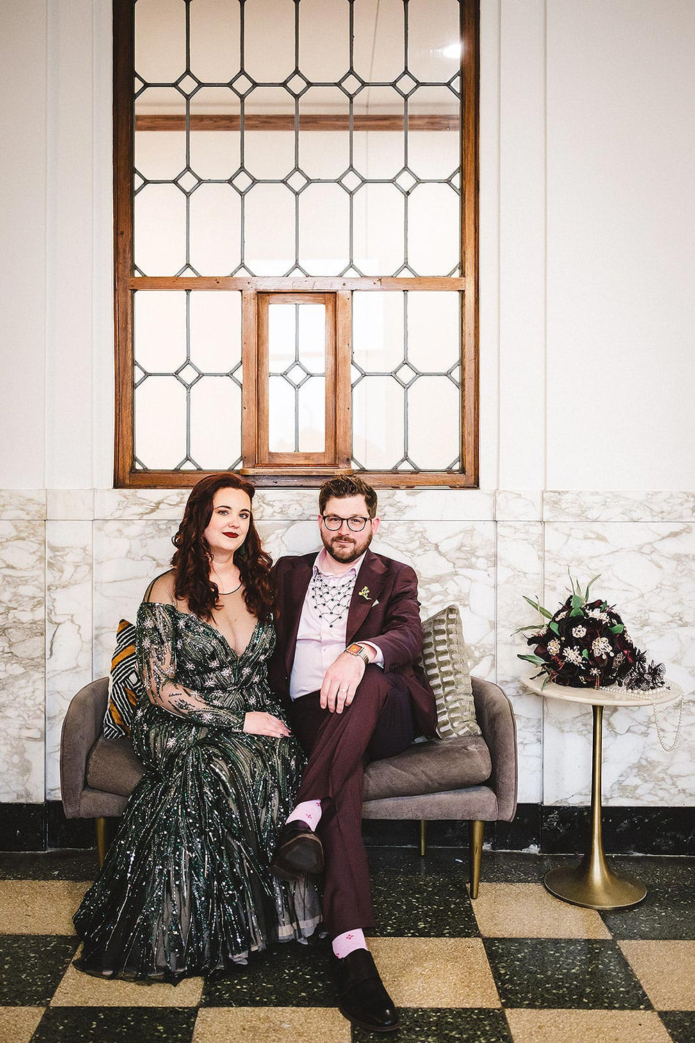 Bride and groom seated on a velvet loveseat in front of a marble wall with a vintage wood-frame window, bride in the Jessica forest green beaded gown by Angela Kim Couture with a dark burgundy bridal bouquet beside them