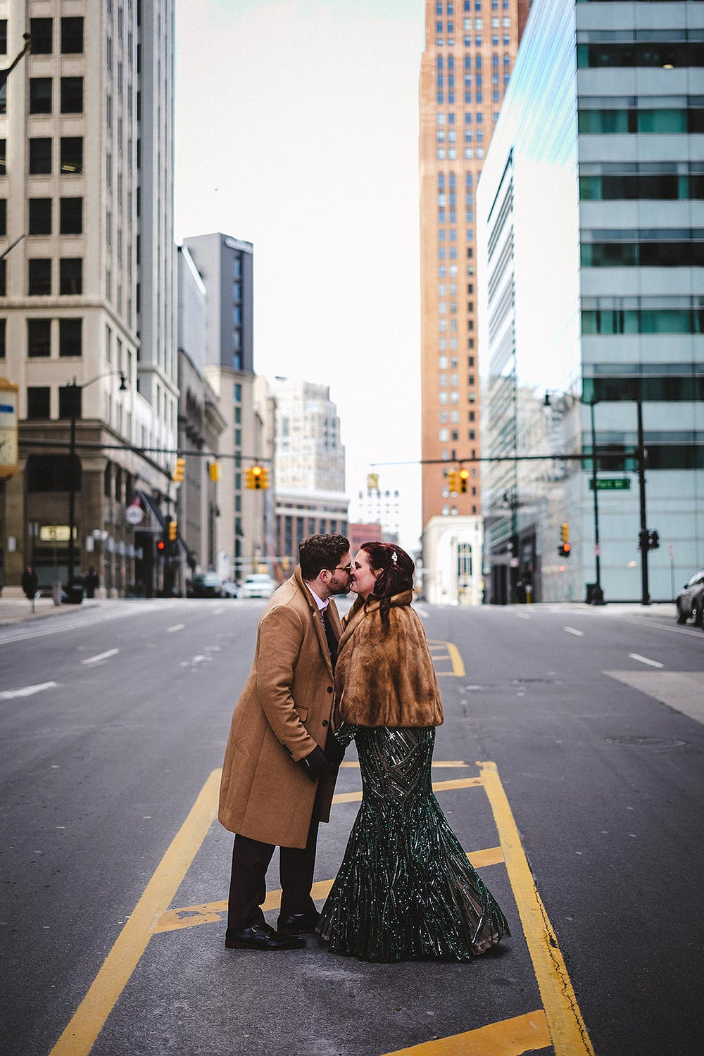 Bride and groom sharing a kiss in the middle of a downtown city intersection in winter, bride in the Jessica forest green beaded wedding gown by Angela Kim Couture with a vintage fur cape, urban skyscraper skyline in the background