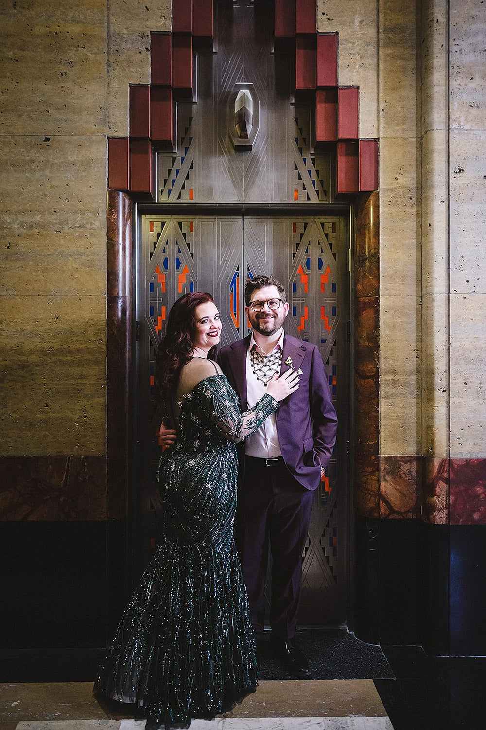 Couple standing in front of an ornate Art Deco elevator door with geometric gold, orange and blue enamel inlay — bride wearing the Jessica forest green trumpet bridal gown by Angela Kim Couture, showcasing the full beaded silhouette
