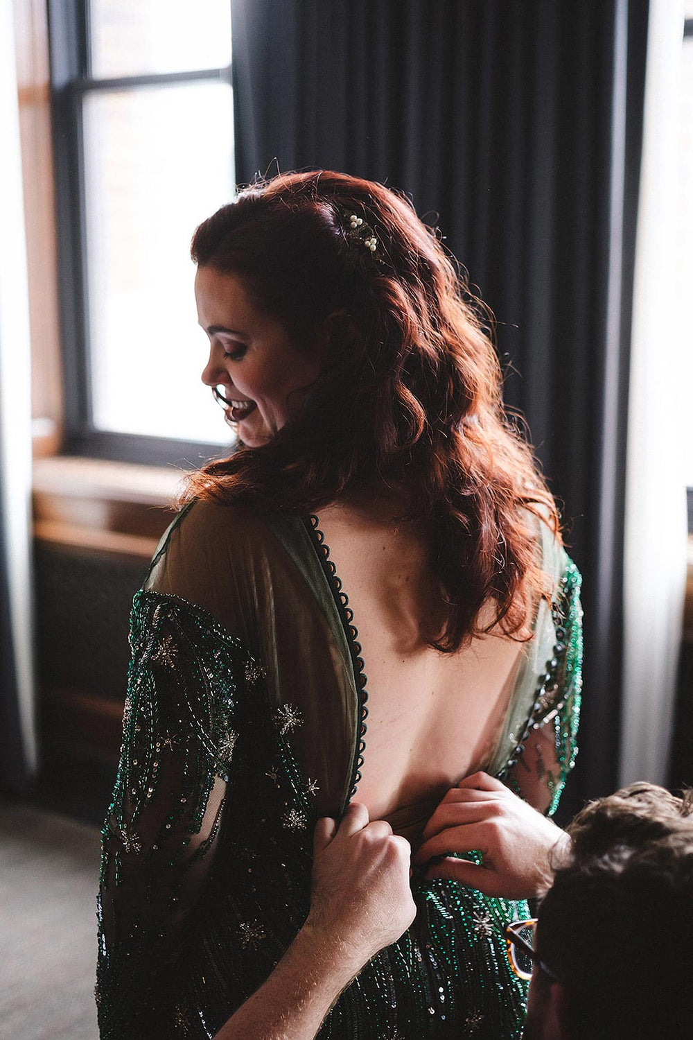 Getting-ready moment showing the groom fastening the illusion tulle open back of the Jessica green wedding dress by Angela Kim Couture, with the crystal and sequin star-motif beaded sleeves in sharp detail