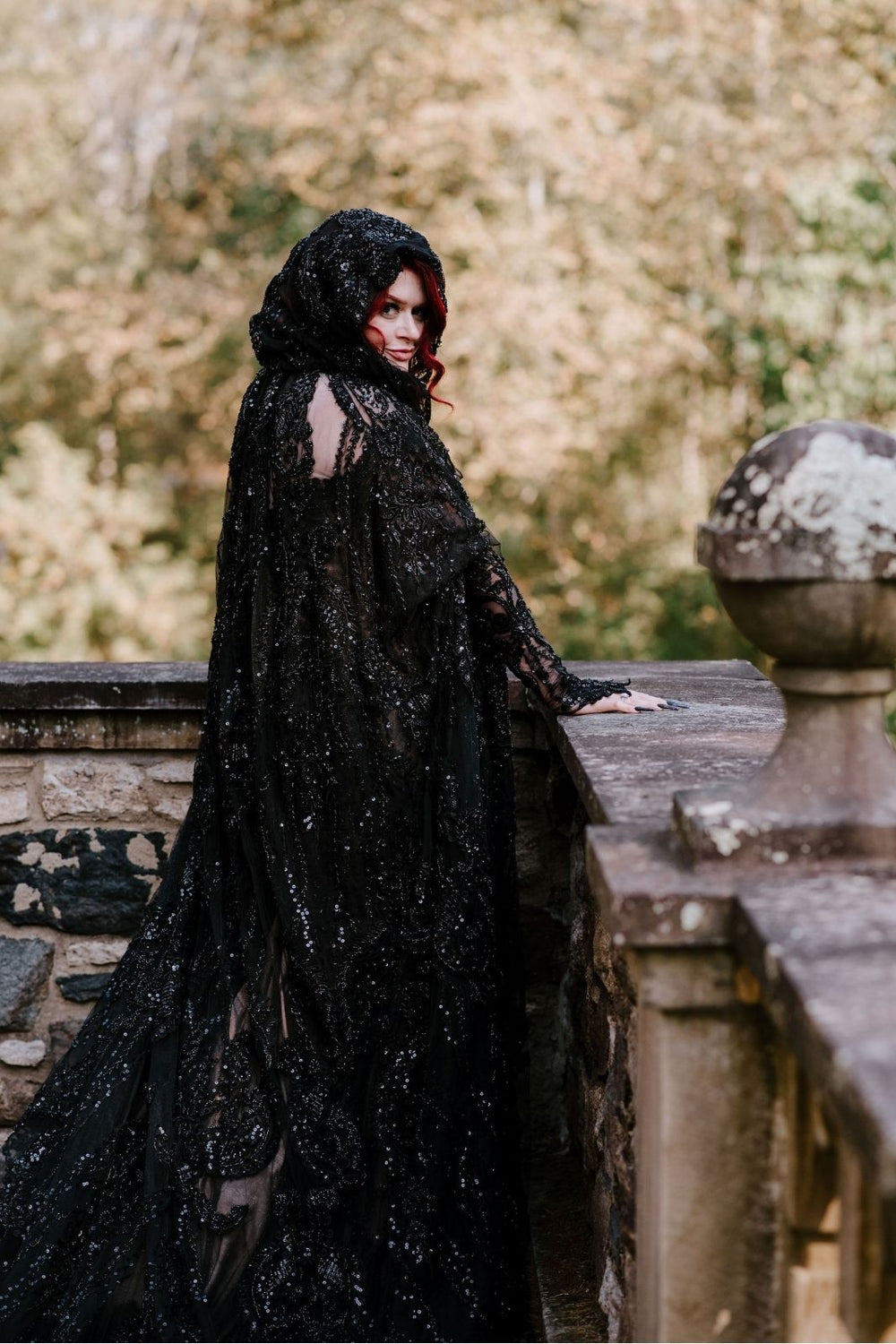 Bride leaning against a stone castle terrace railing — the Kati fit-and-flare black lace beaded dress and a matching cape by Angela Kim Couture draping dramatically with intricate beading pattern and finger-loop sleeve cuff visible