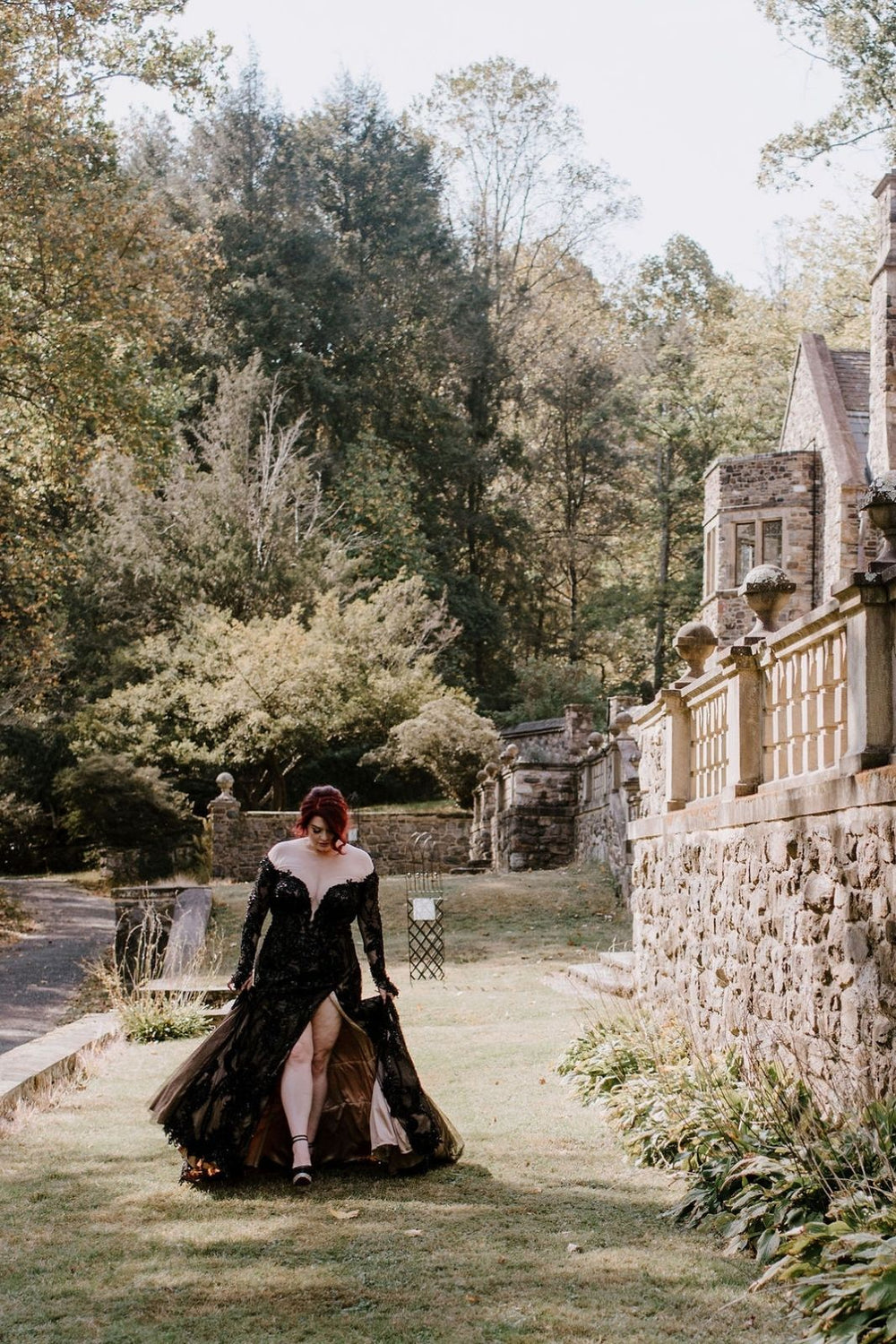 Bride walking alone through a stone castle garden in the Kati black lace fit-and-flare wedding gown by Angela Kim Couture — deep plunging V neckline, thigh-high slit 