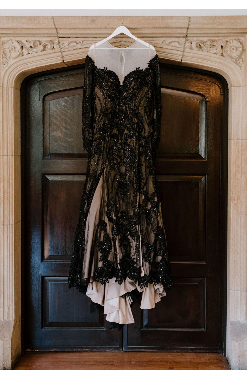 Getting-ready detail of the Kati gothic wedding gown by Angela Kim Couture hanging on a dark wood door — fit-and-flare skirt, black beaded lace over nude satin underlining and the scalloped lace hemline