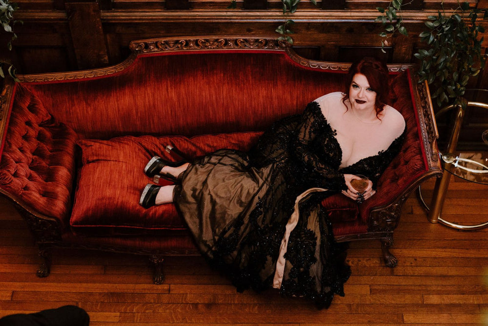 Bride lounging on a vintage burgundy chesterfield sofa in the Kati black lace gothic wedding gown by Angela Kim Couture, long sleeves with finger loops, dark lip, champagne glass, moody wood-paneled interior