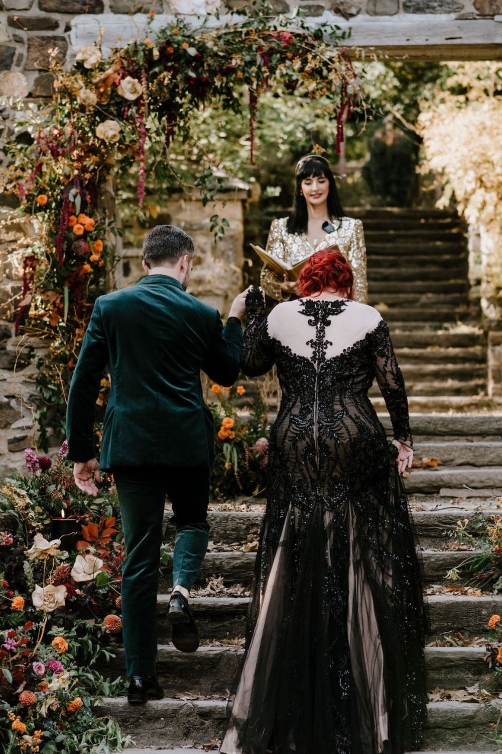Back view of couple ascending floral-lined stone staircase — the Kati gothic gown's scoop back illusion tulle with black lace corset boning by Angela Kim Couture fully visible.