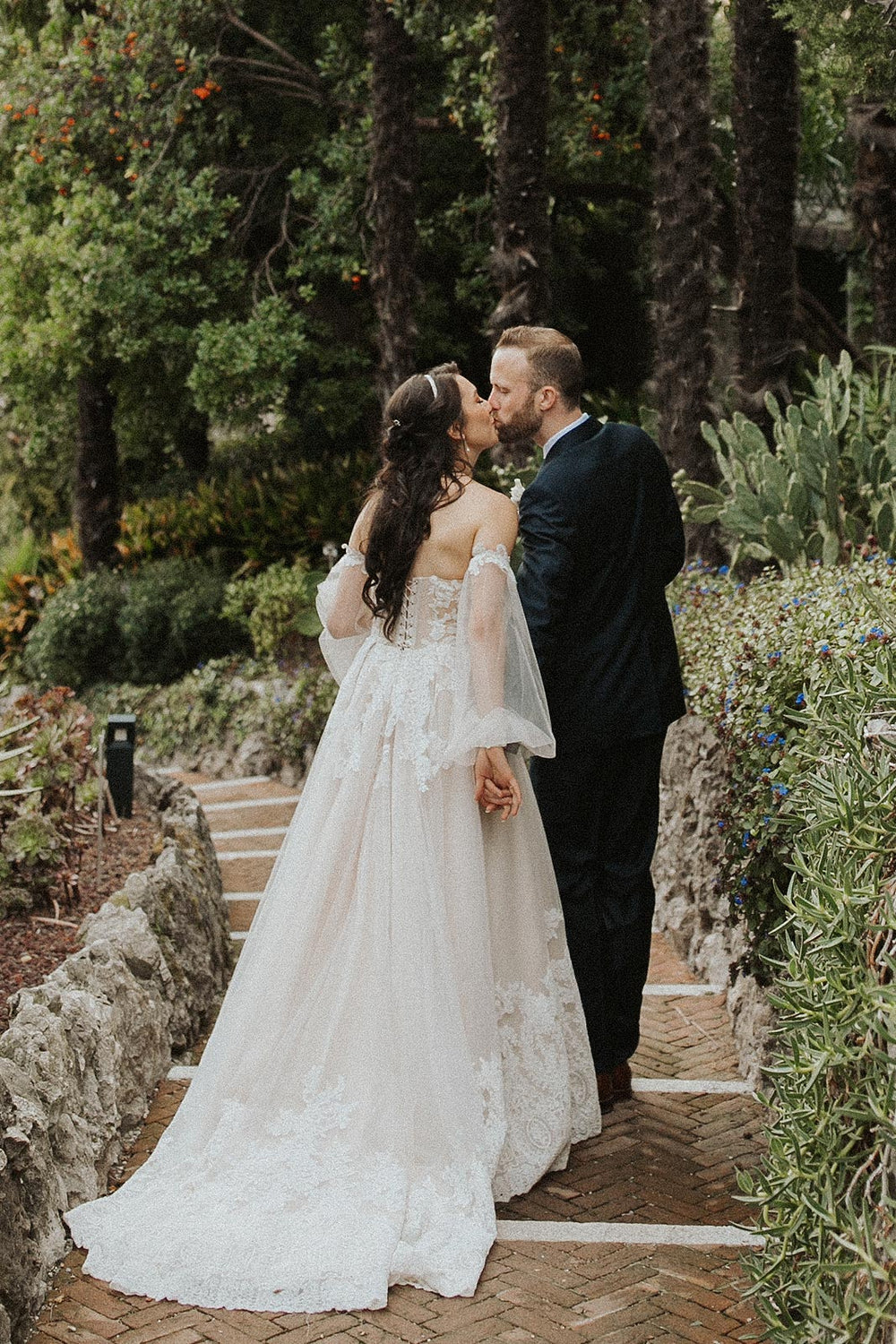 Back view of bride and groom sharing a kiss on Italian garden stone stairs, showcasing the Morgan wedding dress by Angela Kim Couture with bishop sleeves, lace A-line skirt, and corset back