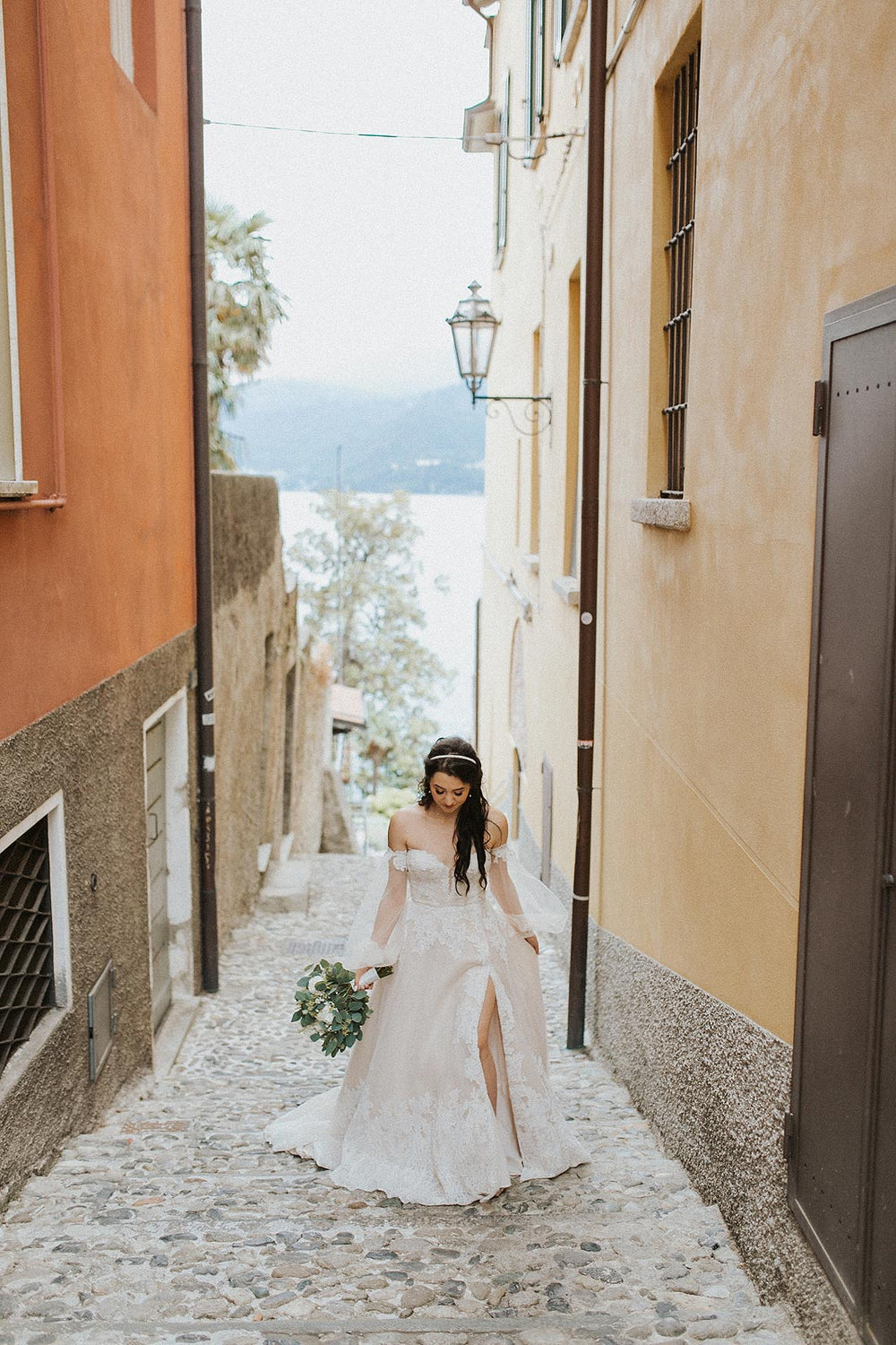 Full-length portrait of bride walking alone through a narrow Italian village cobblestone alley in the Morgan wedding dress by Angela Kim Couture, A-line lace skirt with slit and bishop sleeves, holding a green-and-white bouquet
