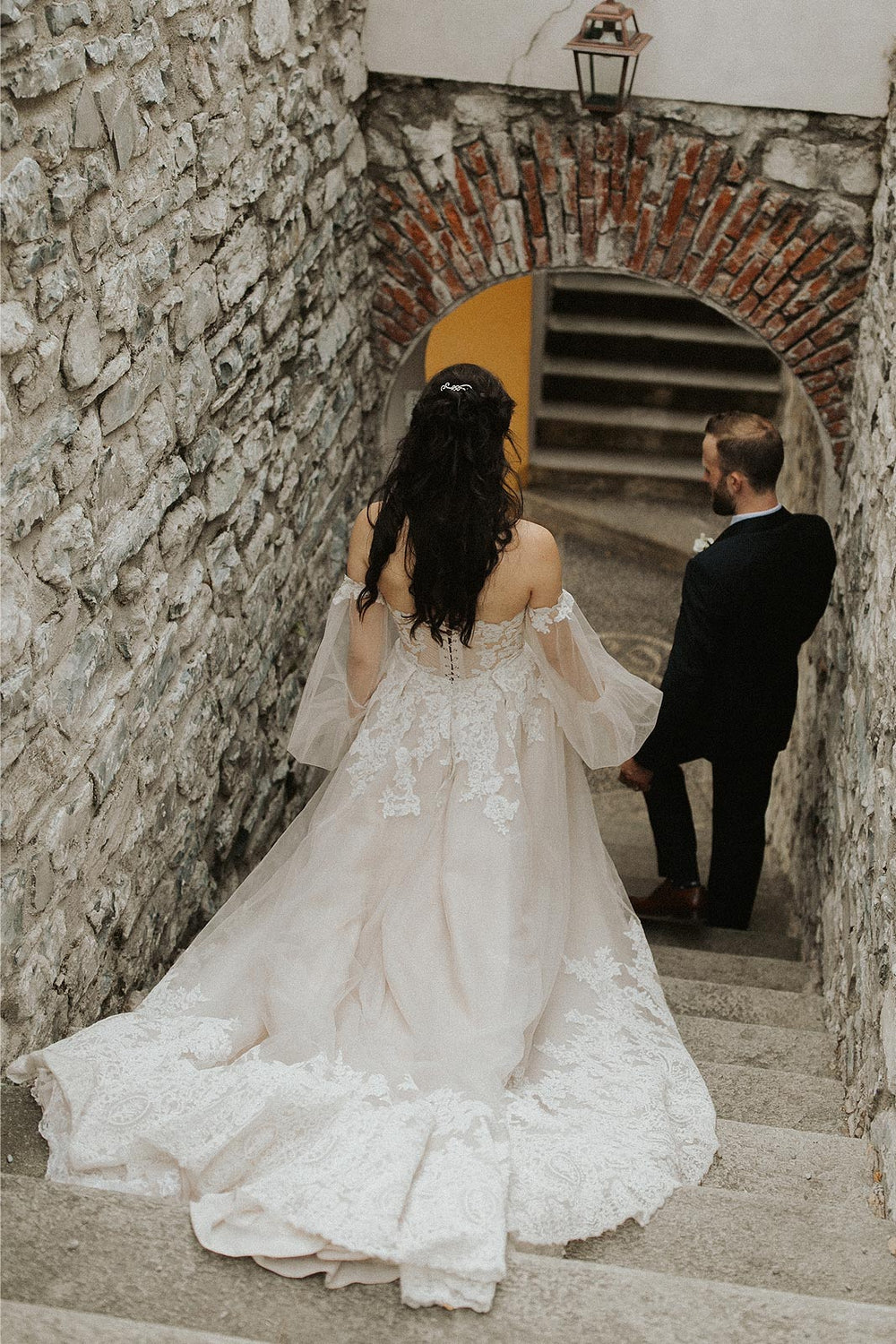 The Morgan couture wedding dress by Angela Kim Couture with lace A-line train sweeping dramatically down ancient Italian stone stairs between rough stone walls, bishop sleeves visible above