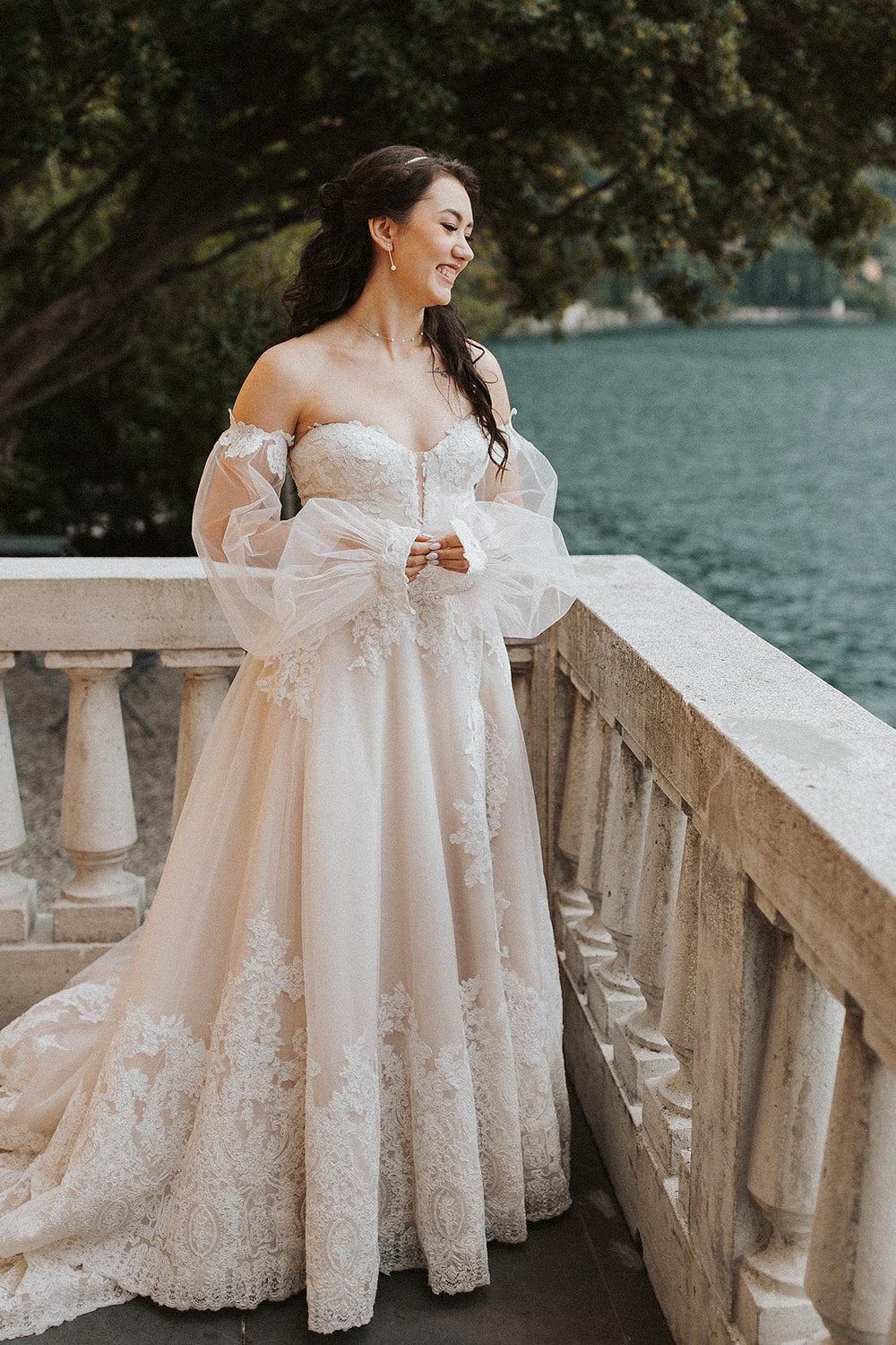 ride wearing the Morgan destination wedding dress by Angela Kim Couture seated on an Italian lakeside terrace, smiling in the sheer off-shoulder bishop sleeves and lace corset bodice