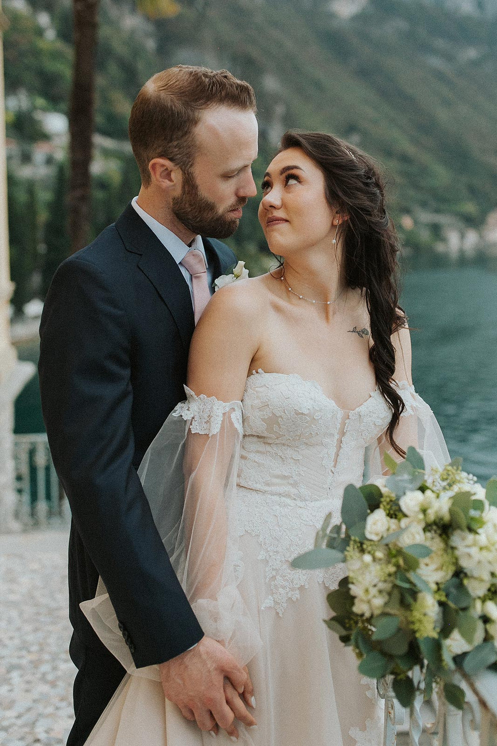 Half-body portrait of bride holding a white and green bouquet by an Italian lake wearing the Morgan couture wedding dress by Angela Kim Couture, highlighting the sweetheart bodice and sheer off-shoulder bishop sleeves