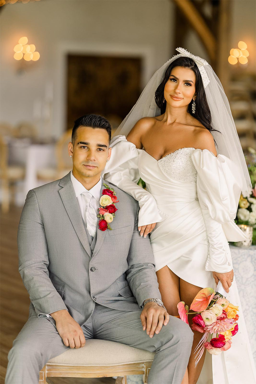Seated couple portrait at an outdoor wedding ceremony with the Veronica mini wedding dress by Angela Kim Couture, showing the short form-fitting skirt, Juliet sleeve cuff, and colorful tropical bouquet beside the groom in a light grey suit