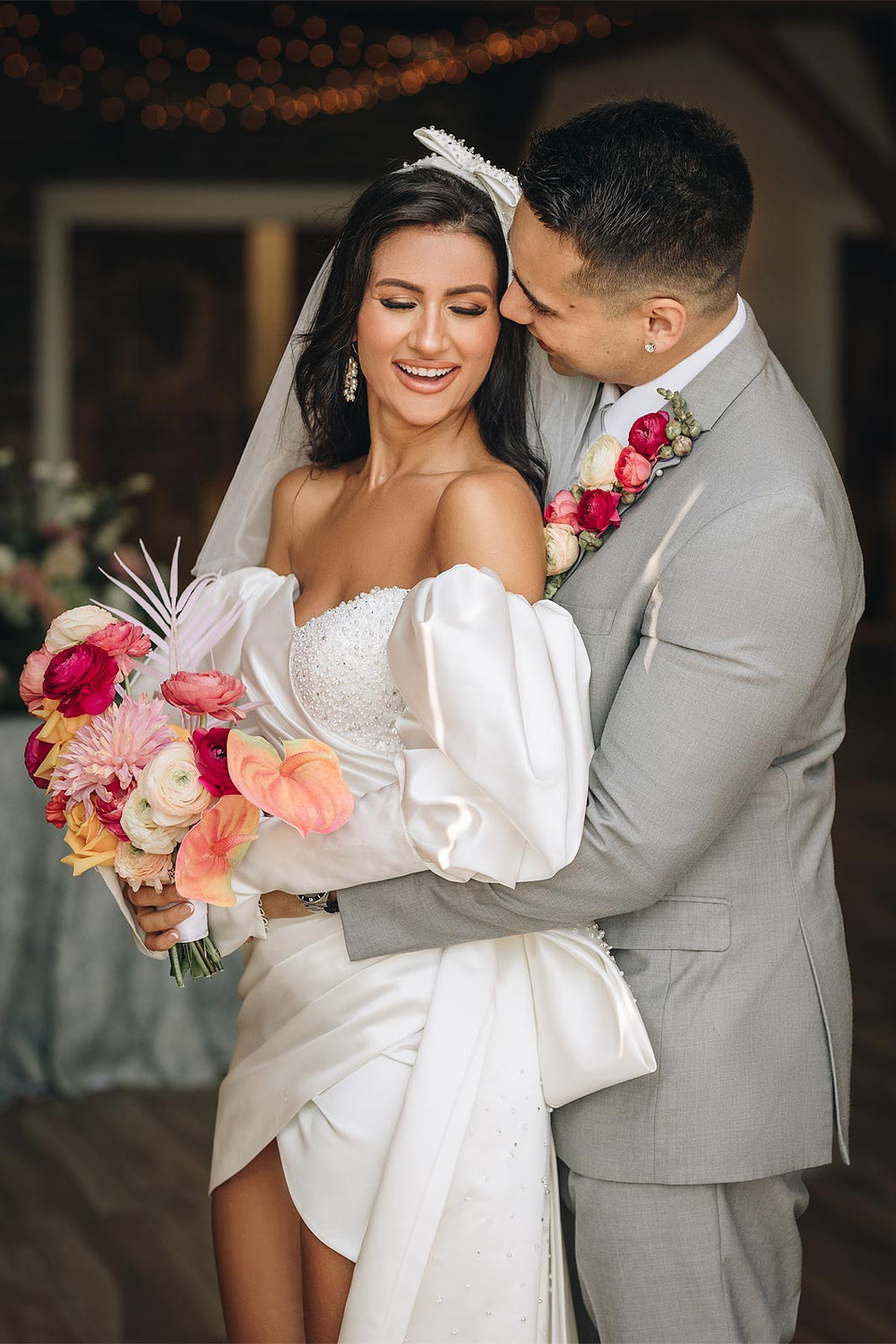 Bride and groom embracing in the Veronica short beaded wedding dress by Angela Kim Couture, Juliet sleeves draped over the groom's arms, crystal-pearl beaded bodice visible, with a vibrant coral and pink tropical bouquet