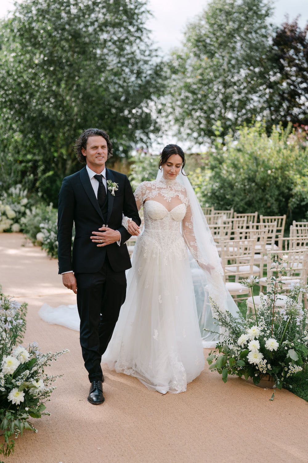 Bride Blaire and groom walking through an English garden during their wedding ceremony, showcasing the full sweeping overskirt of her custom three-piece Angela Kim Couture bridal ensemble