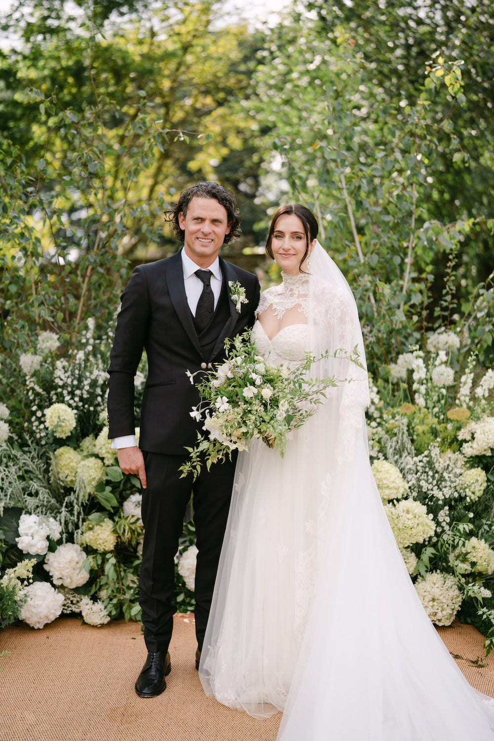 ceremony shot of Blaire and her groom at the altar surrounded by a lush white floral arch, showing the dramatic train of her custom overskirt wedding gown by Angela Kim Couture
