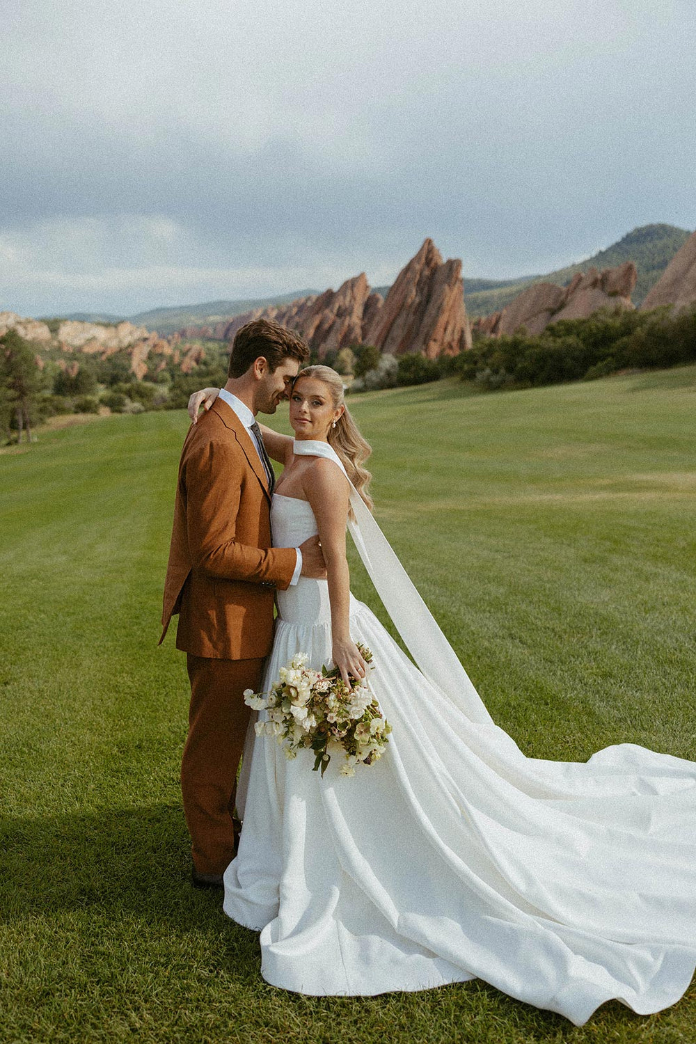 Hannah and her groom standing on green grass, showcasing the full spread of her custom crepe wedding gown train and delicate neck veil designed by Angela Kim Couture