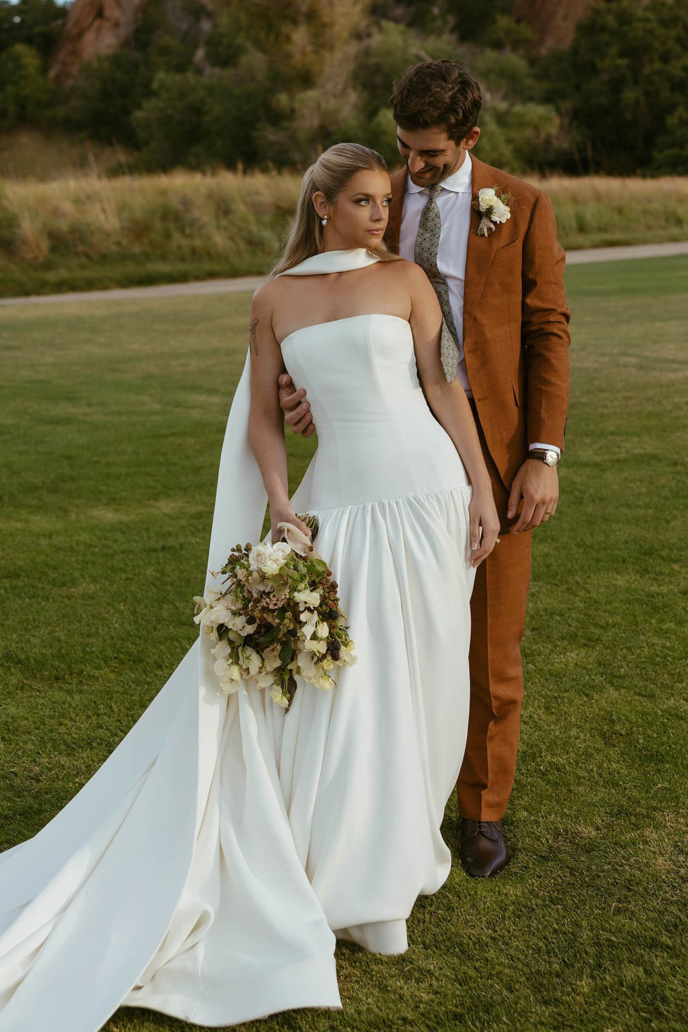 Bride Hannah and her groom in an embrace outdoors, Hannah wearing a custom strapless heavy-weight crepe wedding dress with a dramatic draped silk neck bow designed by Angela Kim Couture, red rock mountains in the background