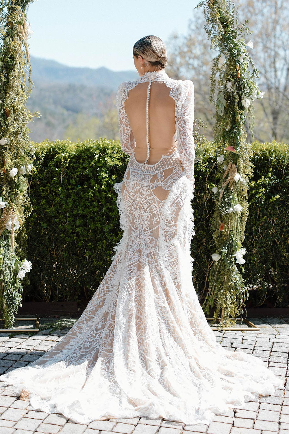 Full-length back view of Peyton's custom lace wedding gown by Angela Kim Couture showing the dramatic open back with delicate button closure, fit-and-flare silhouette and train on cobblestone, green floral arch visible