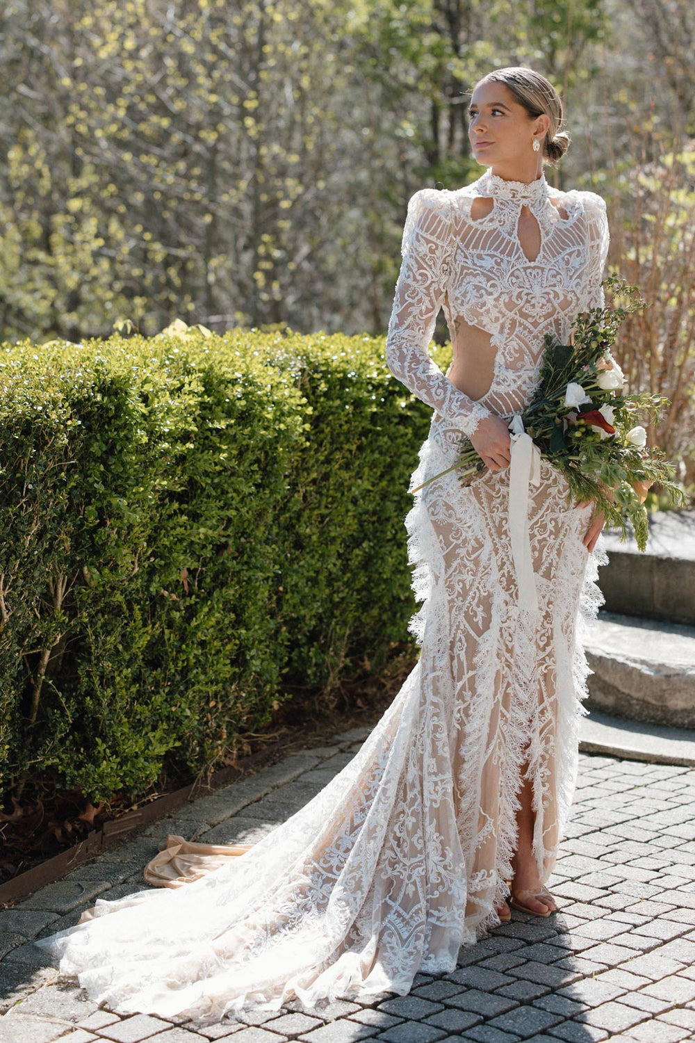 Bride Peyton in a custom high-neck intricate lace fit-and-flare wedding gown with keyhole cut-out neckline and subtle puff sleeves, holding a wildflower bouquet, designed by Angela Kim Couture at an outdoor garden venue with mountain views