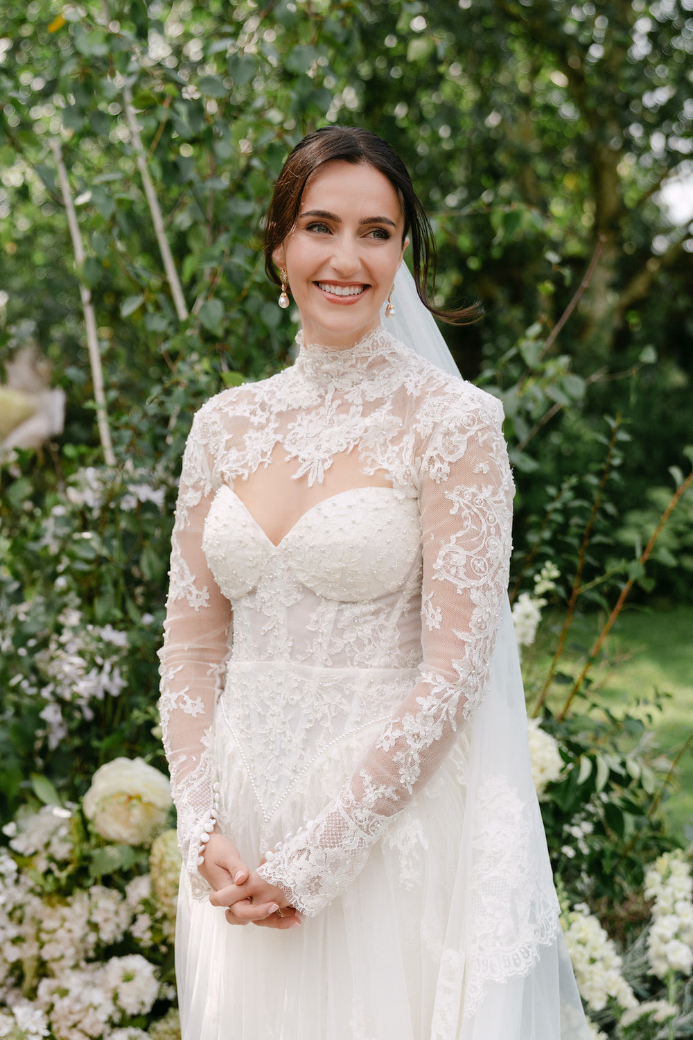 Angela Kim Couture bride Blaire smiling in a custom high-neck sheer lace bolero wedding gown with pearl embellishments photographed in an outdoor garden setting