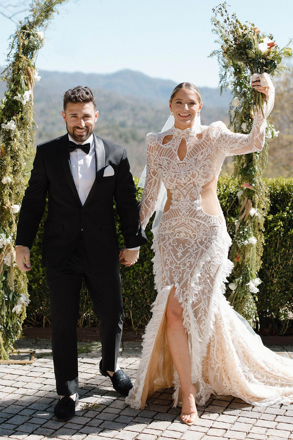 Bride Peyton and her groom holding wildflower bouquets together at their outdoor ceremony with a sweeping mountain backdrop and green floral arch columns, Peyton in her custom lace wedding dress by Angela Kim Couture